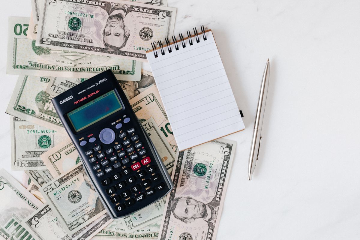 US dollar banknotes beside a calculator, blank notepad and pen.