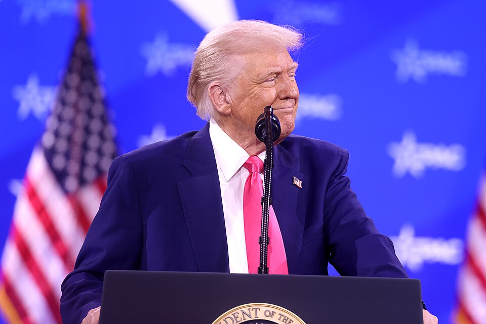 Donald Trump in a dark suit and red tie at a microphone, with a blue backdrop and a U.S. flag behind him.