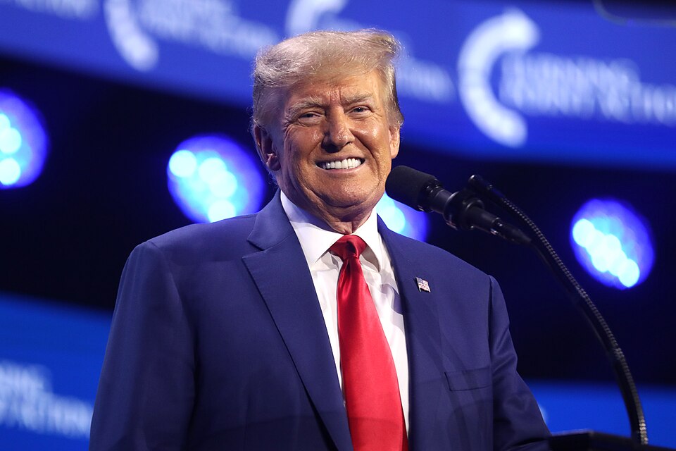 Archive photograph of Donald Trump in a blue suit and red tie, smiling beside a microphone.