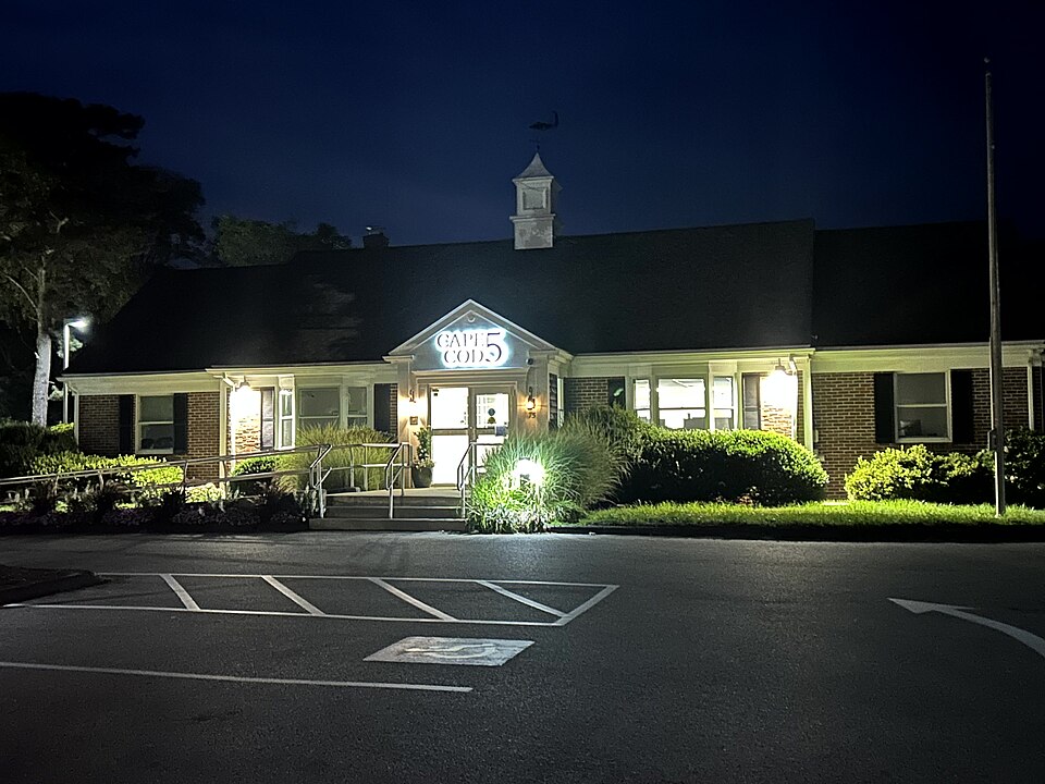 A Cape Cod Five bank building has an illuminated entrance beside an empty parking area at night.