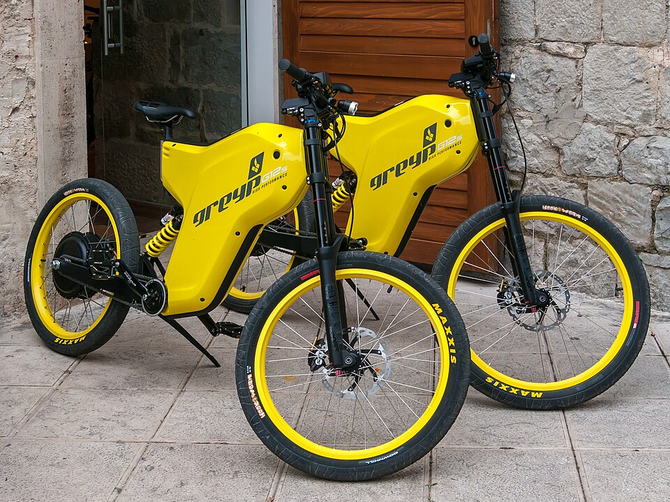 Two yellow electric bicycles stand on paving outside a stone building.