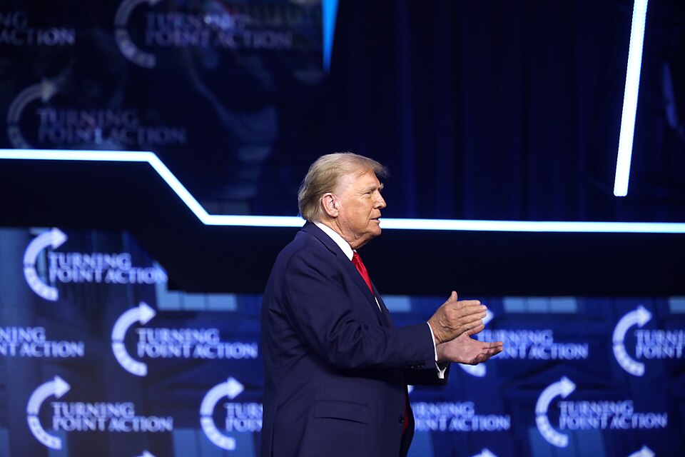 Donald Trump in a dark suit and red tie, clapping on a stage with Turning Point Action signs.