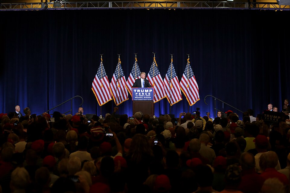Donald Trump stands behind a Trump-Pence campaign lectern, with American flags behind him and an audience in the foreground.