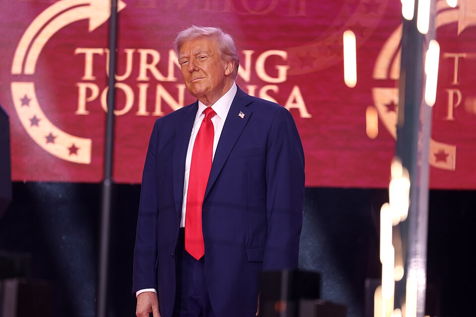 Donald Trump in a dark suit and red tie standing before a Turning Point USA backdrop.
