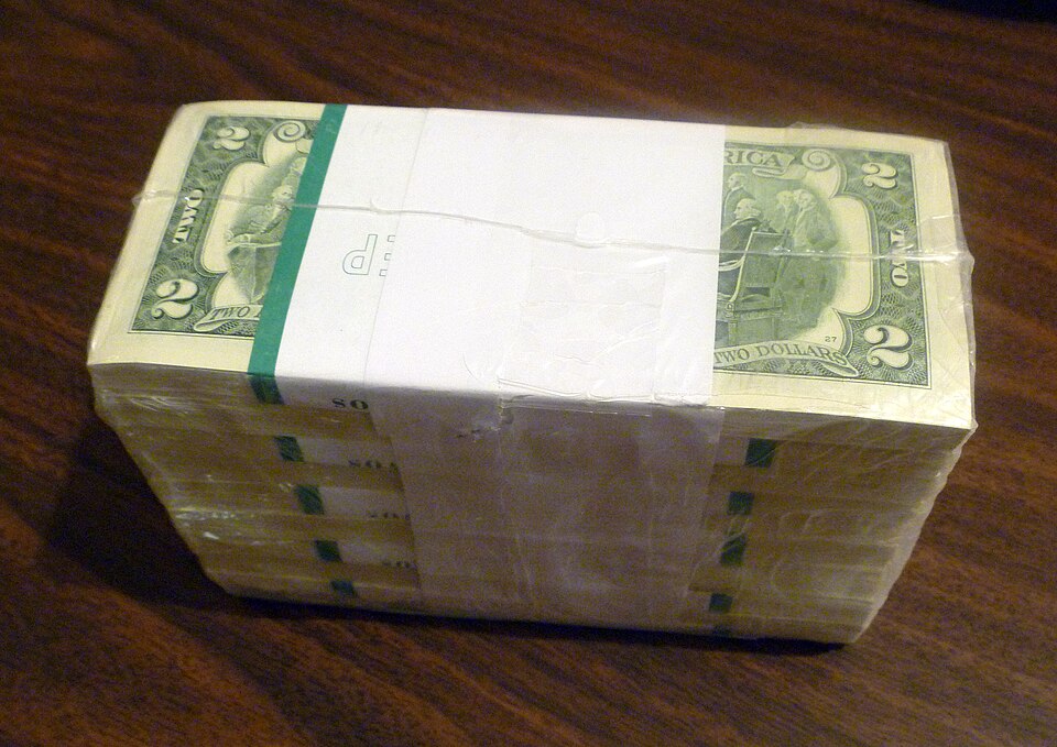 A shrink-wrapped stack of U.S. two-dollar bills on a wooden surface.