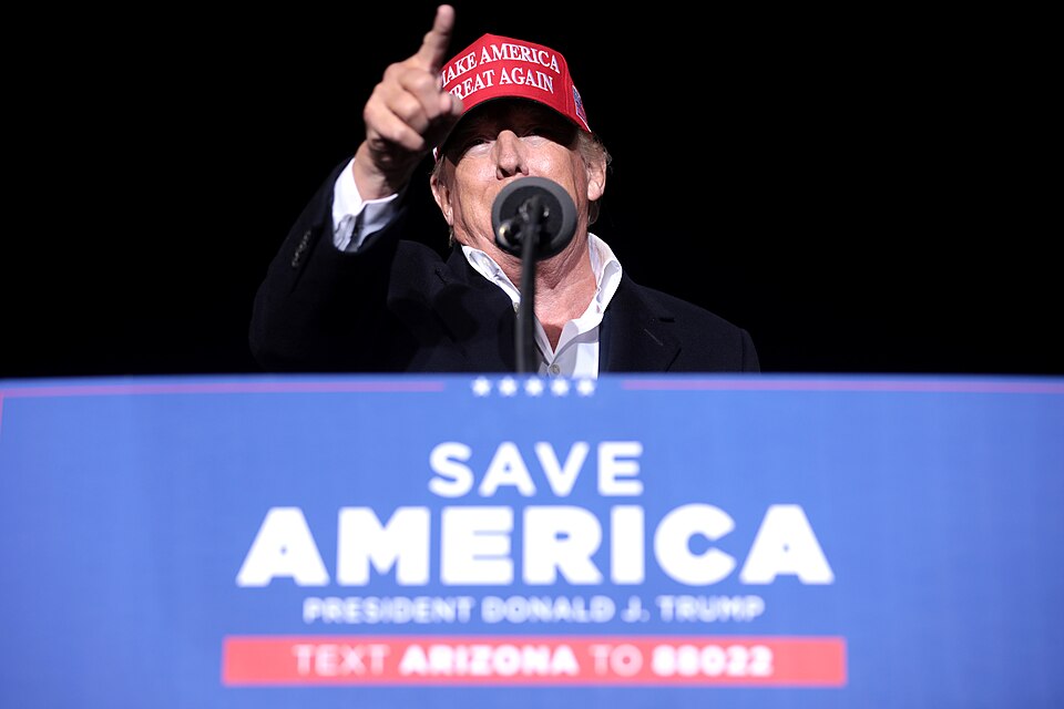 Donald Trump wears a red campaign cap and gestures while speaking into a microphone behind a Save America sign.