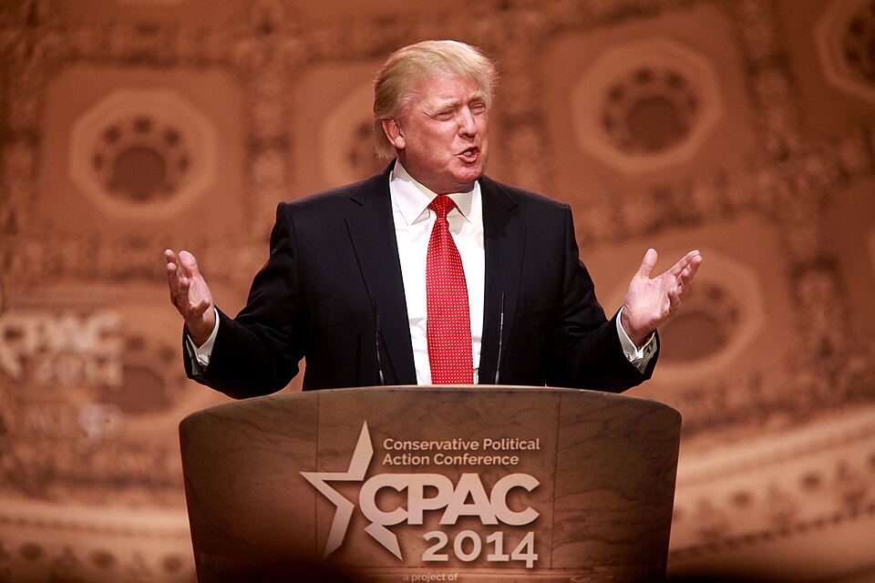 Donald Trump gestures with both hands while standing behind a CPAC 2014 lectern.