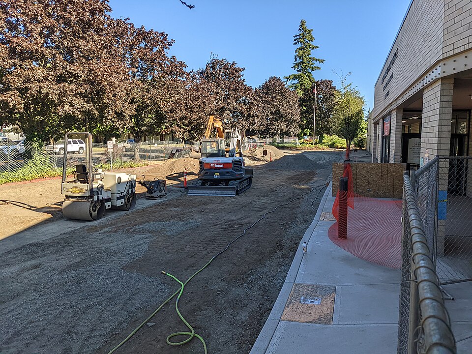 Construction equipment and an unpaved area outside a Post Office entrance, with trees and temporary fencing nearby.