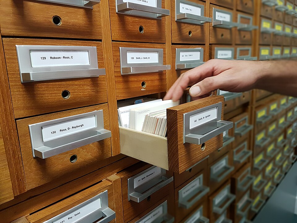 A hand reaches into an open card drawer among rows of labeled wooden filing drawers.