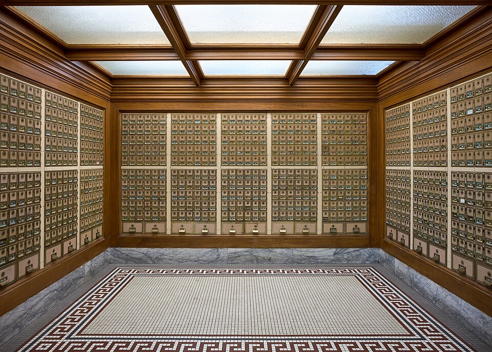 Rows of individual metal PO boxes line three walls of a wood-trimmed post office lobby with a tiled floor.