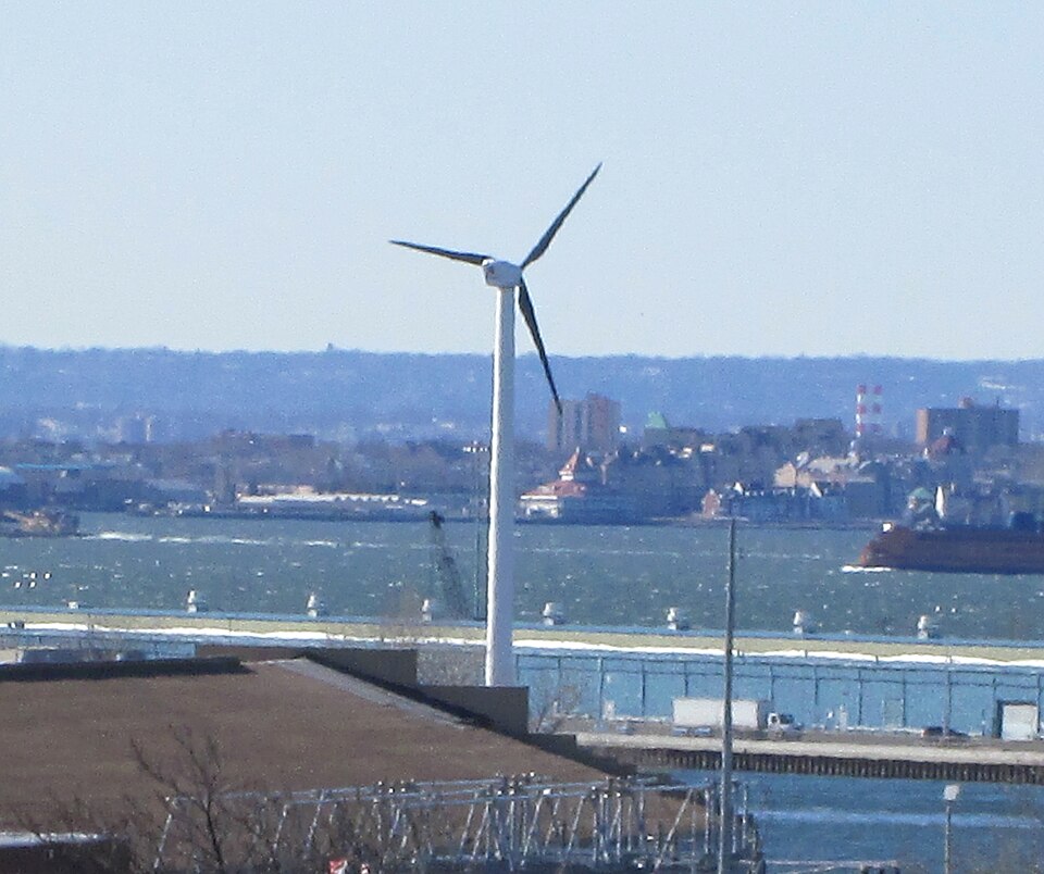 A white wind turbine rises above waterfront buildings, with water and ships behind it.