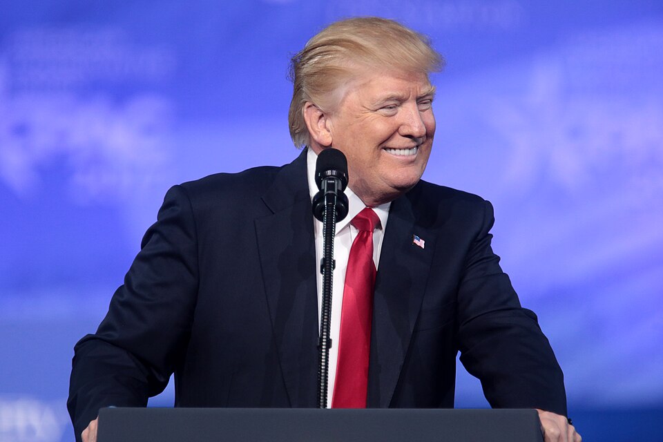 Donald Trump stands at a lectern with a microphone against a blue background.
