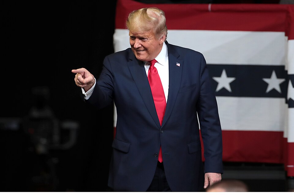 Donald Trump smiles and points while standing in front of a red, white and blue backdrop.