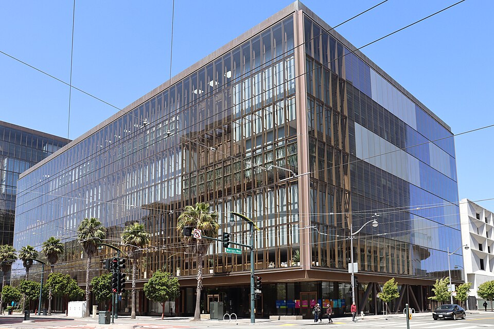 Glass-fronted office building at a street intersection, with palm trees and overhead wires.
