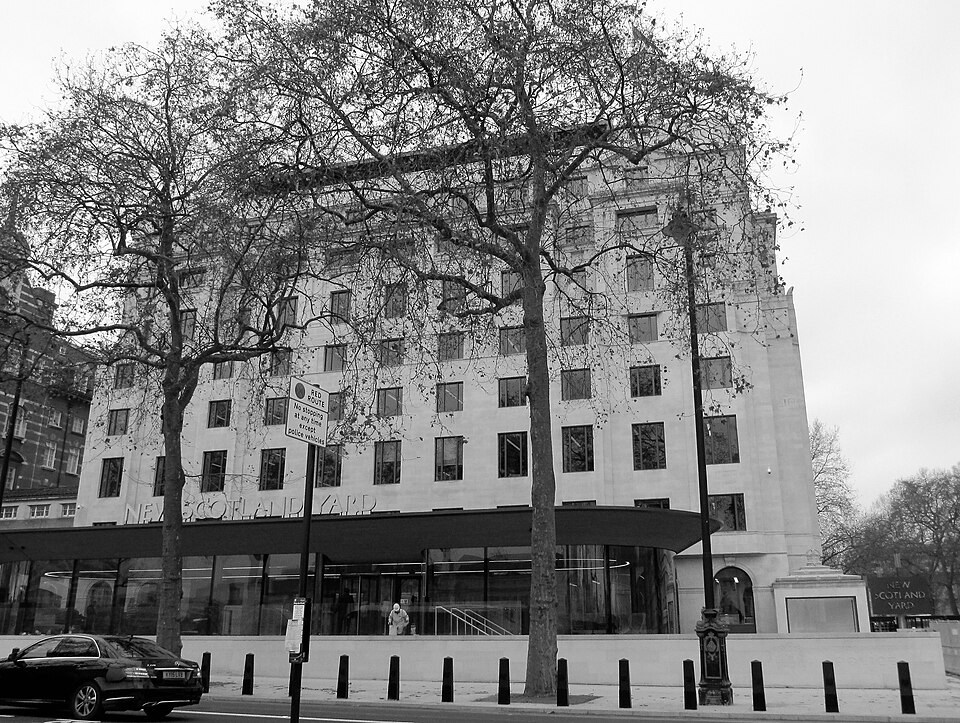 Black-and-white exterior view of New Scotland Yard behind trees, with a car on the street.
