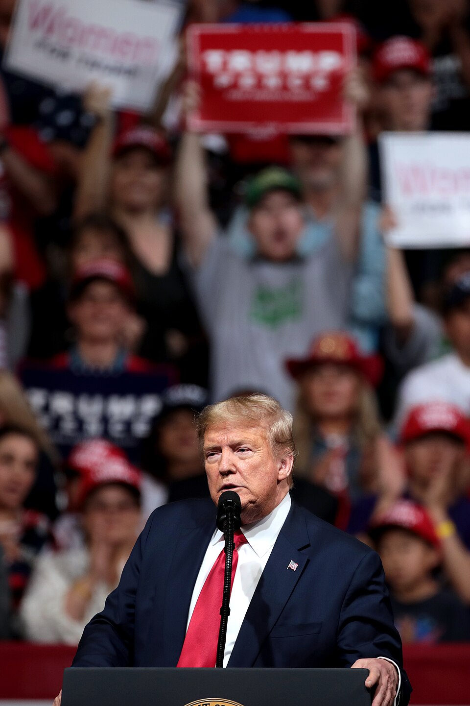 Donald Trump in a dark suit and red tie at a microphone, with supporters holding signs behind him.