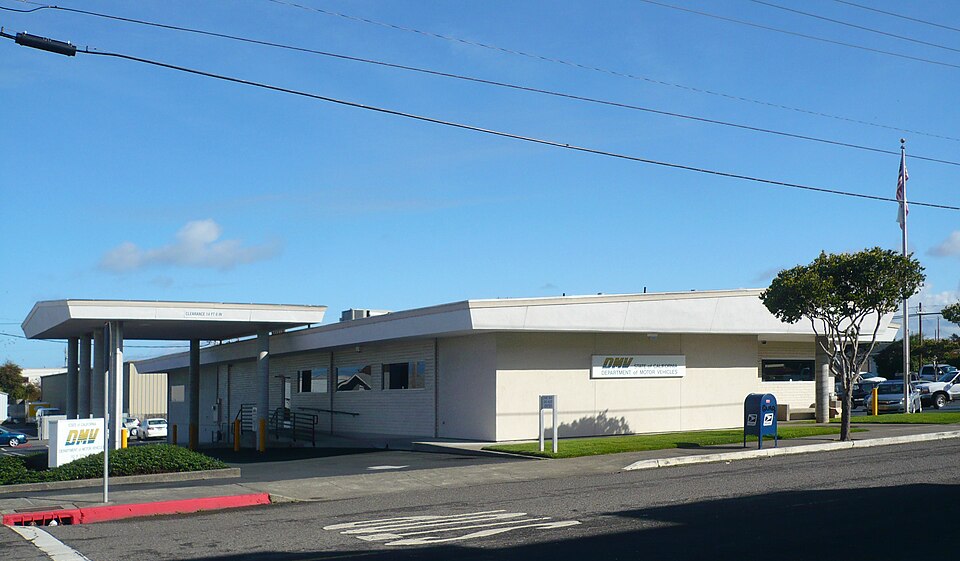 A low, light-colored DMV office building with a covered entrance and a blue mailbox beside the street.