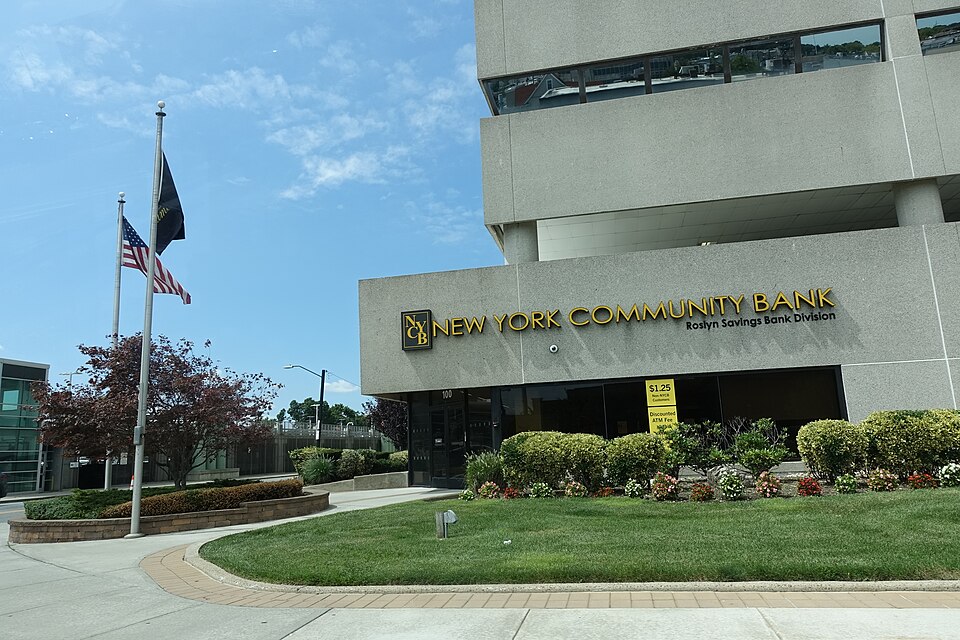 A bank building has a New York Community Bank sign above its entrance, with shrubs, a lawn and flagpoles outside.