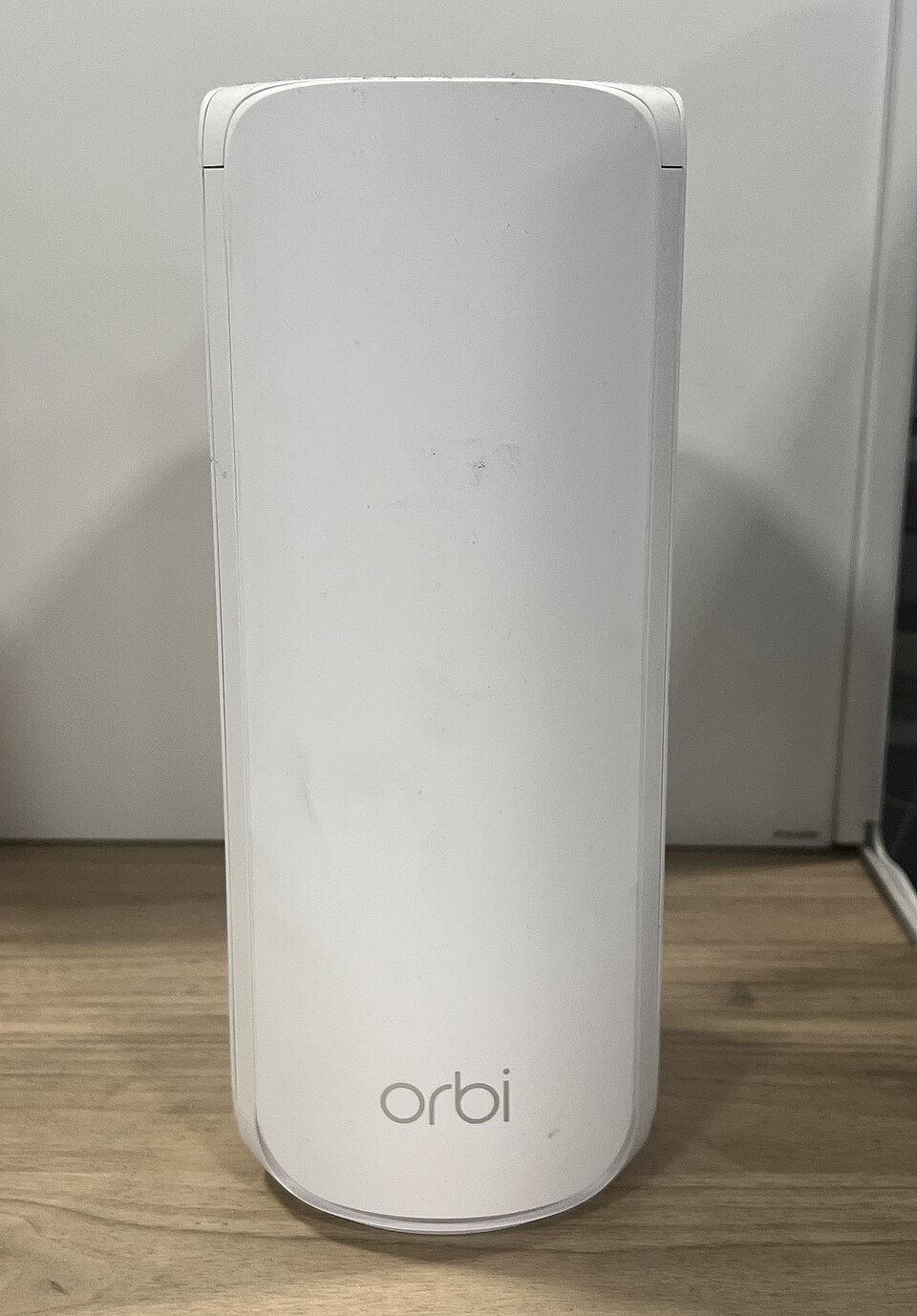A tall white Orbi router stands on a wooden surface against a light-colored wall.