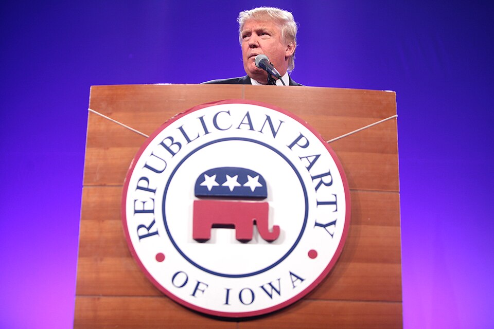 Donald Trump stands behind a wooden lectern bearing a Republican Party of Iowa emblem, with a microphone and purple backdrop.