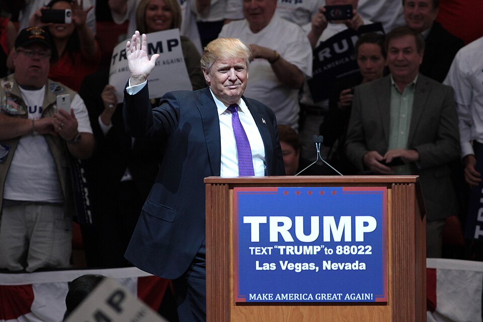 Donald Trump raises one hand beside a campaign lectern, with supporters and campaign signs behind him.