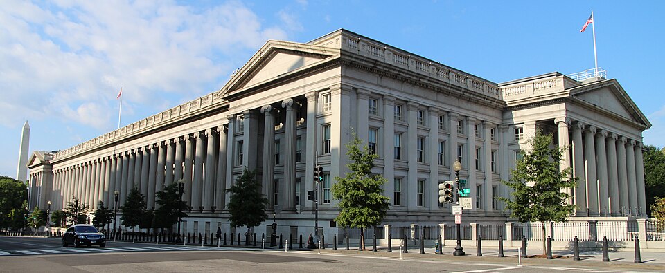 Archive view of the U.S. Treasury building, with stone columns, U.S. flags and a street in front.