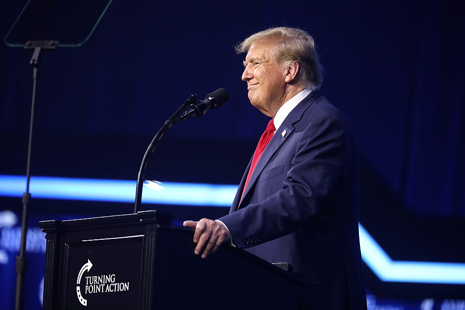 Donald Trump in a dark suit and red tie stands at a lectern with a microphone against a dark blue background.