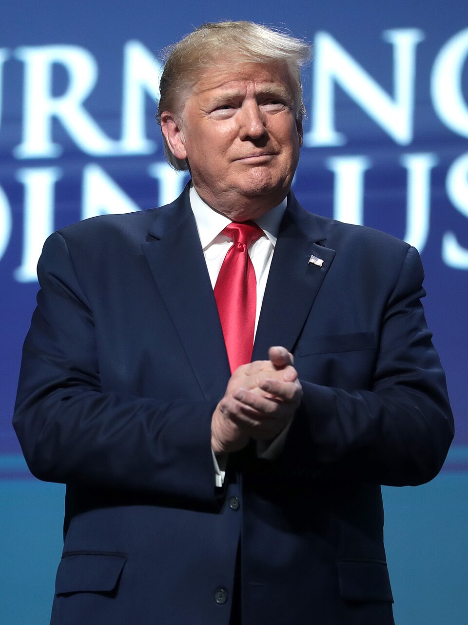 Donald Trump wearing a dark suit and red tie, with his hands together in front of a blue backdrop.