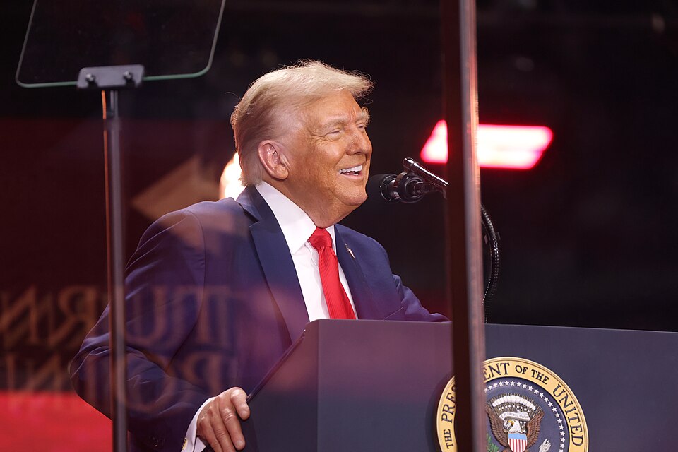 Donald Trump smiles beside microphones at a lectern bearing the presidential seal, with teleprompters in front of him.