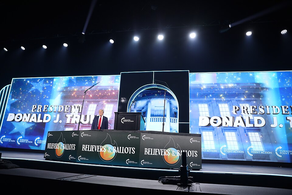 Donald Trump stands on a stage beside a lectern with Believers & Ballots signs and large screens behind him.