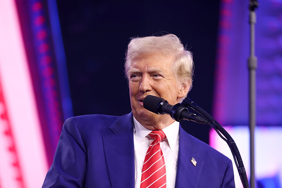 Donald Trump in a blue suit and red striped tie at a microphone against a purple-lit background.