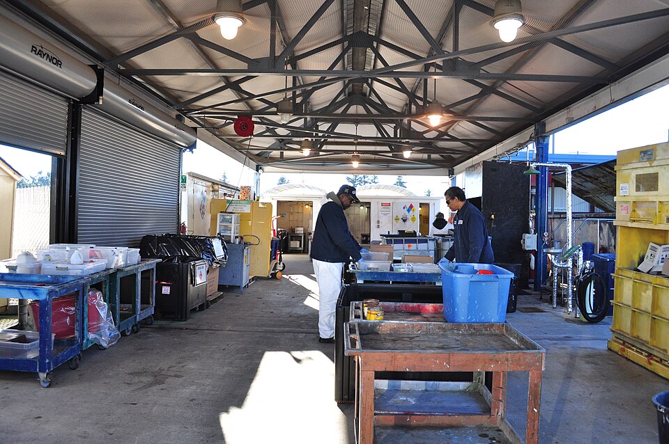 Two people stand beside worktables and containers under a metal roof at a collection facility.