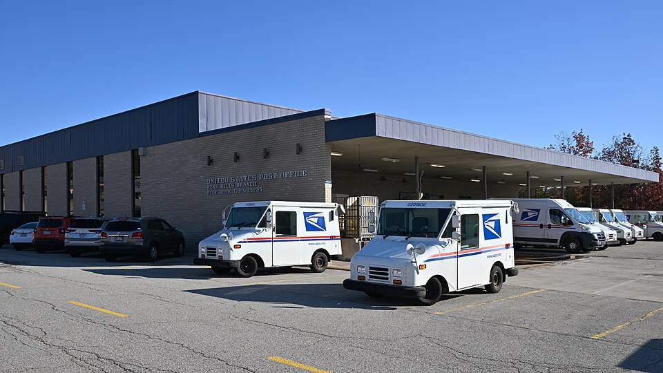White USPS delivery vehicles are parked outside a brick building bearing a United States Post Office sign.